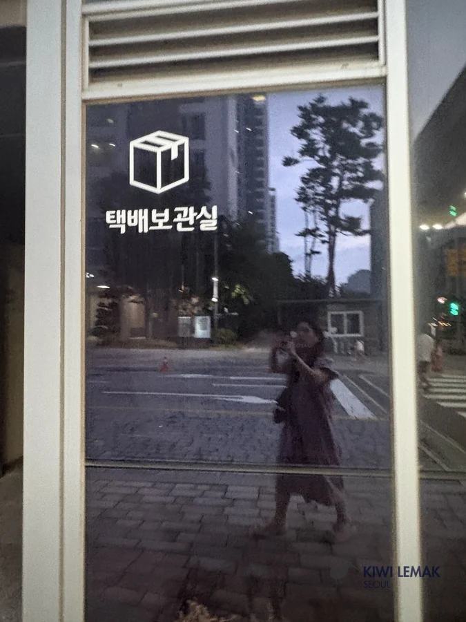 Parcel storage room signage in a Korean apartment building lobby, seen through a window at dusk