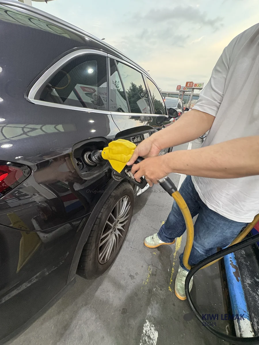 A driver, shown from behind, holds the pump nozzle in the car's open fuel door while fueling, with the rest-stop station visible in the background.