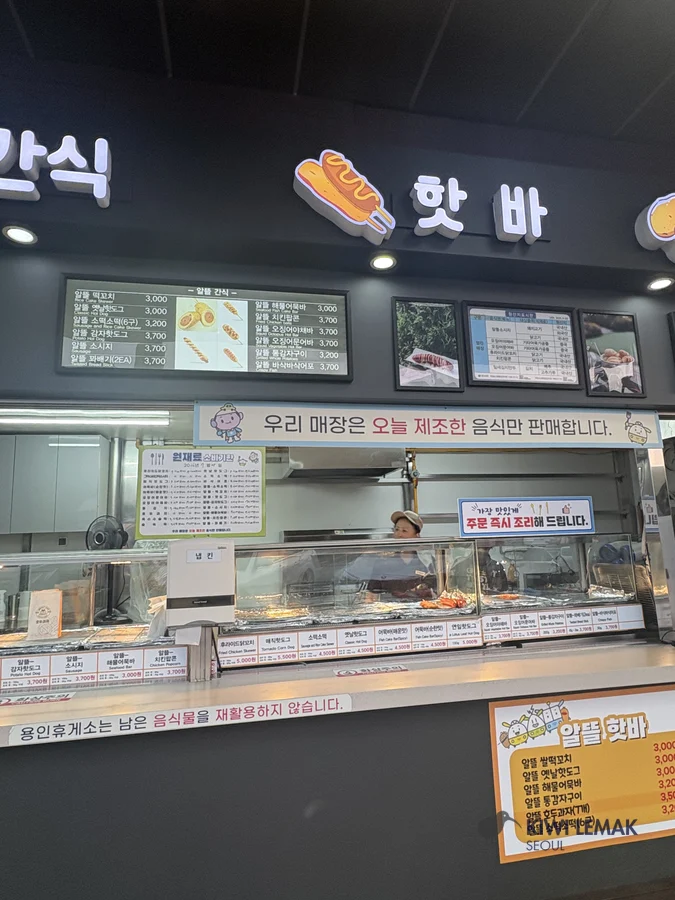 A 'hot bar' snack counter's illuminated menu board lists corn dogs and similar snacks with prices in won, with a staff member visible behind the counter.