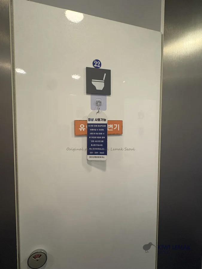 A restroom stall door displays a hanging card reading 'currently available for normal use,' with instructions to flip it to report a malfunction and a facility contact number.